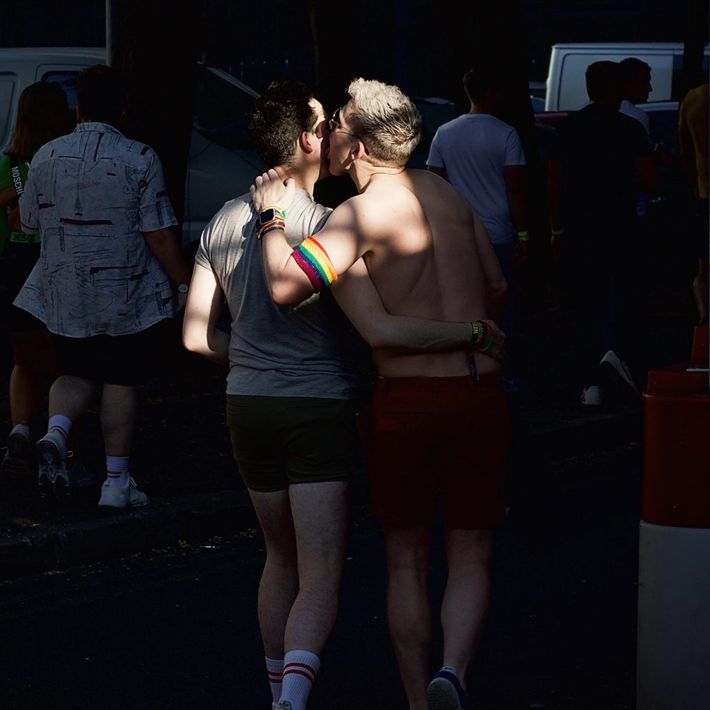 Showing the backs of two men, one with a rainbow armband who have their arms linked around each other