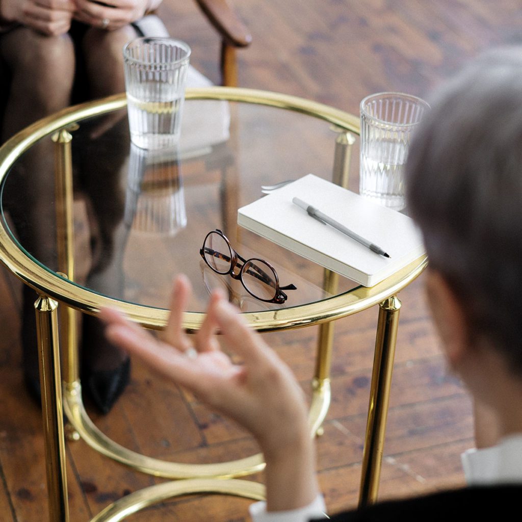 a round glass table with glasses of water on it and a glimpse of two people talking across the table